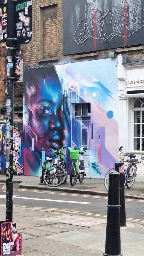 Vibrant street art mural of a face in blue and purple hues on brick building with parked bicycles in urban setting
