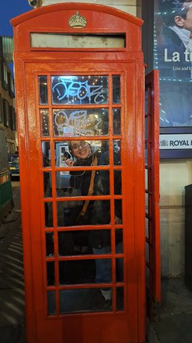 Woman smiling inside iconic red British phone booth with crown symbol and glass panels in urban setting