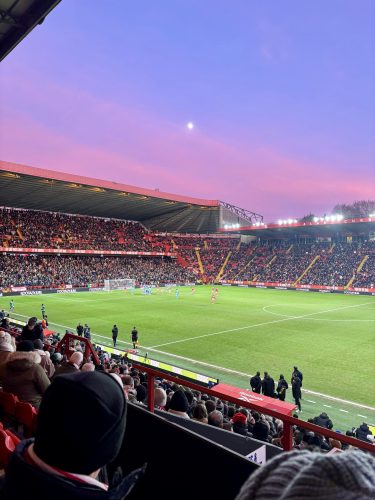 Football stadium at sunset with purple sky, packed red stands, players on green pitch during evening match