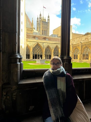 Woman in scarf and glasses sitting by Gothic cathedral cloister window with ornate stone arches and courtyard view
