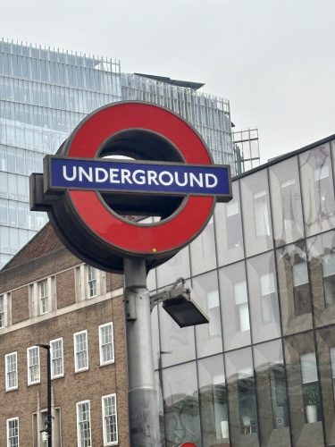 London Underground sign with red circle and blue bar against backdrop of modern glass building and historic brick architecture